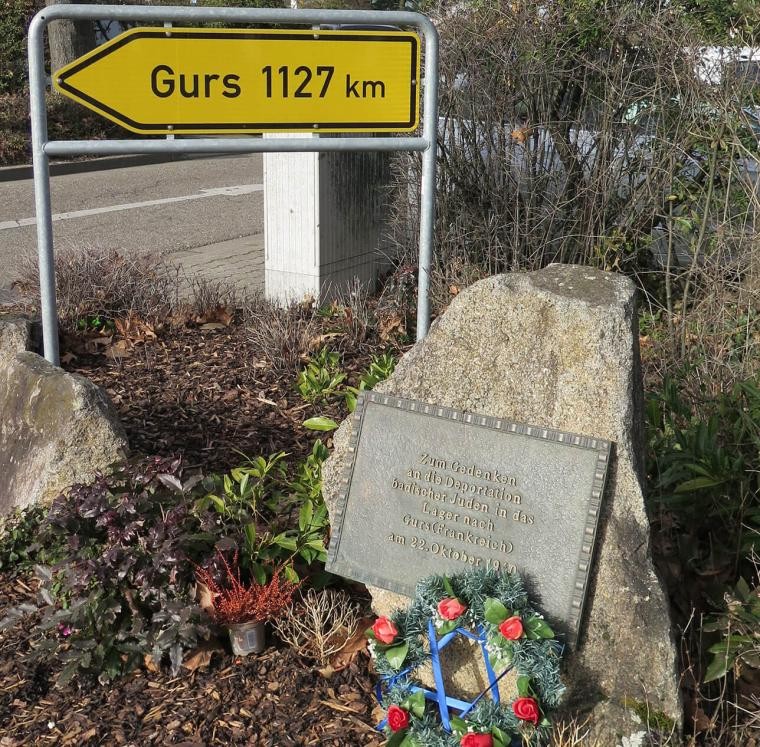Straßenschild mit "Gurs 1127 km" und einem Gedenkstein und Blumen vorn einem Stein