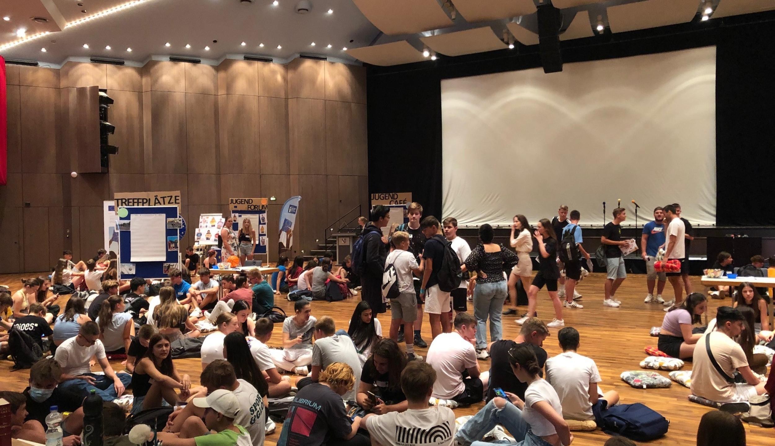Many young people sit and stand in a large hall. There are info stands and a big screen in the background.