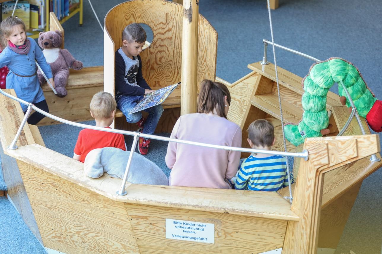 Mehrere Kinder und eine Mutter sitzen auf einer Holzbank, die einem Boot nachempfunden ist und lesen Bücher.