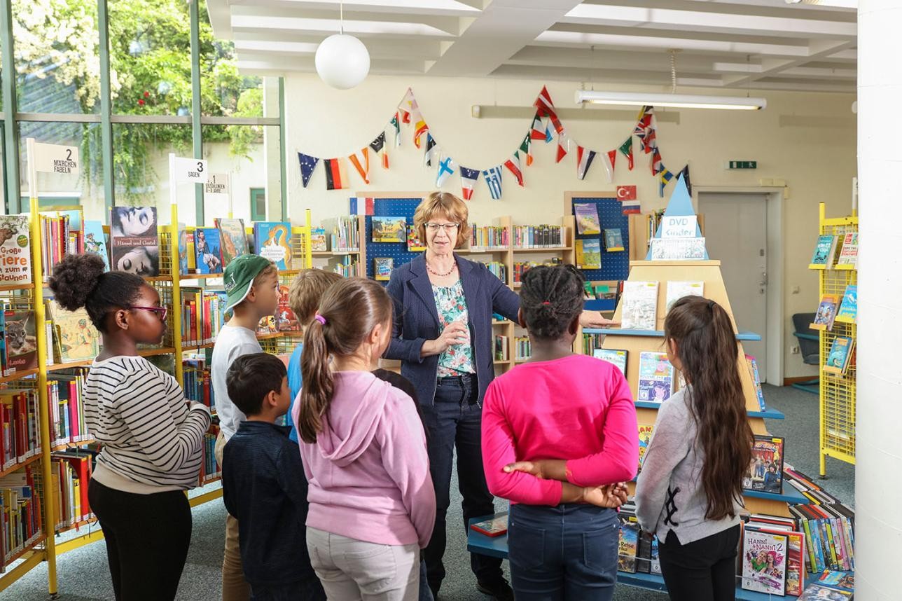 Library tour - several children stand around Birgitt Stellmach while she tells them something. Bookshelves in the background.