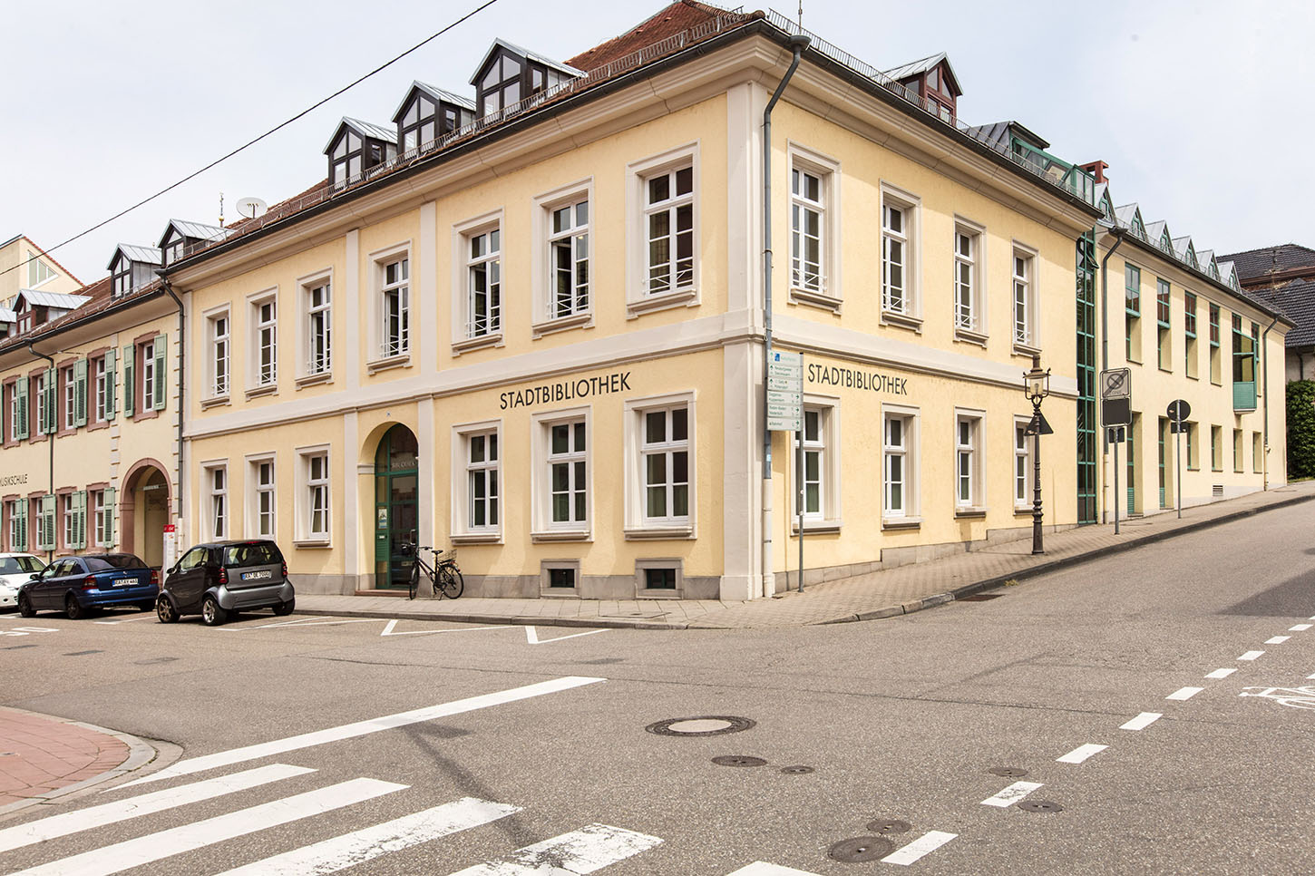 City library exterior shot with intersection, cars parked on the street.